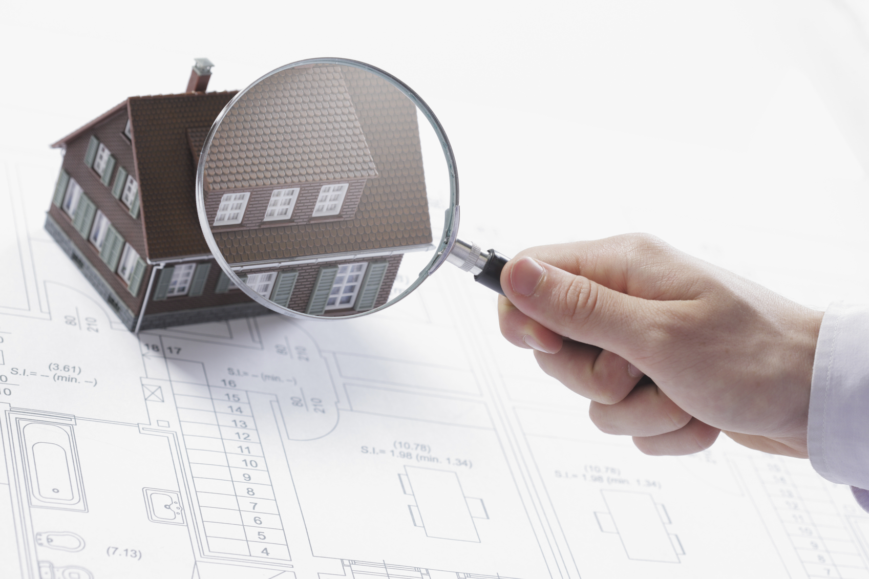Concept image of a home inspection. A male hand holds a magnifying glass over a miniature house.